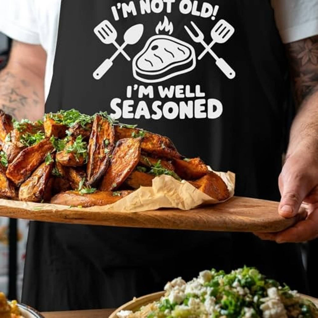 Black apron featuring "I'm not old, I'm well seasoned" grilling graphic over cooked food