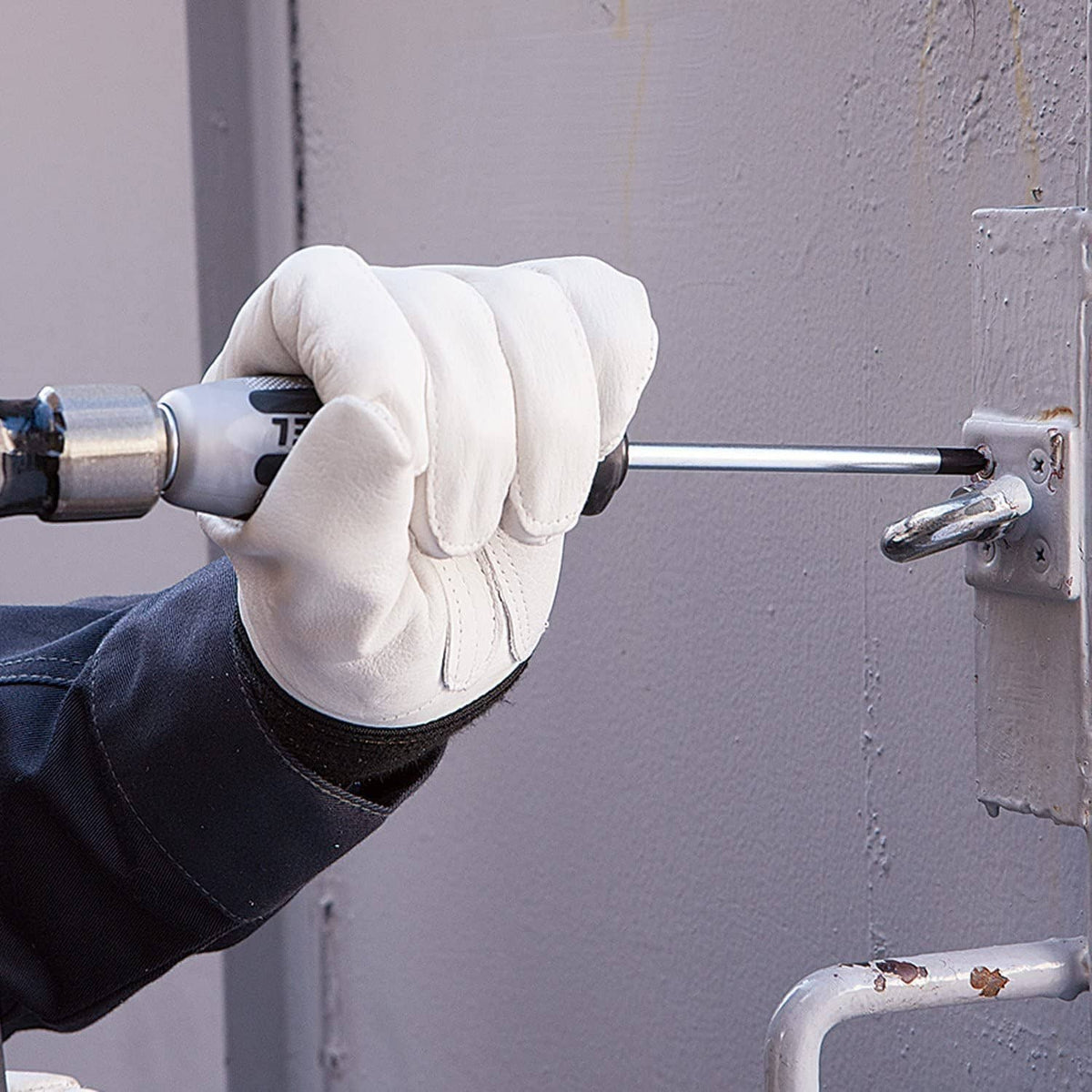 Gloved hand holding impact cross-point screwdriver driving a screw into a panel.