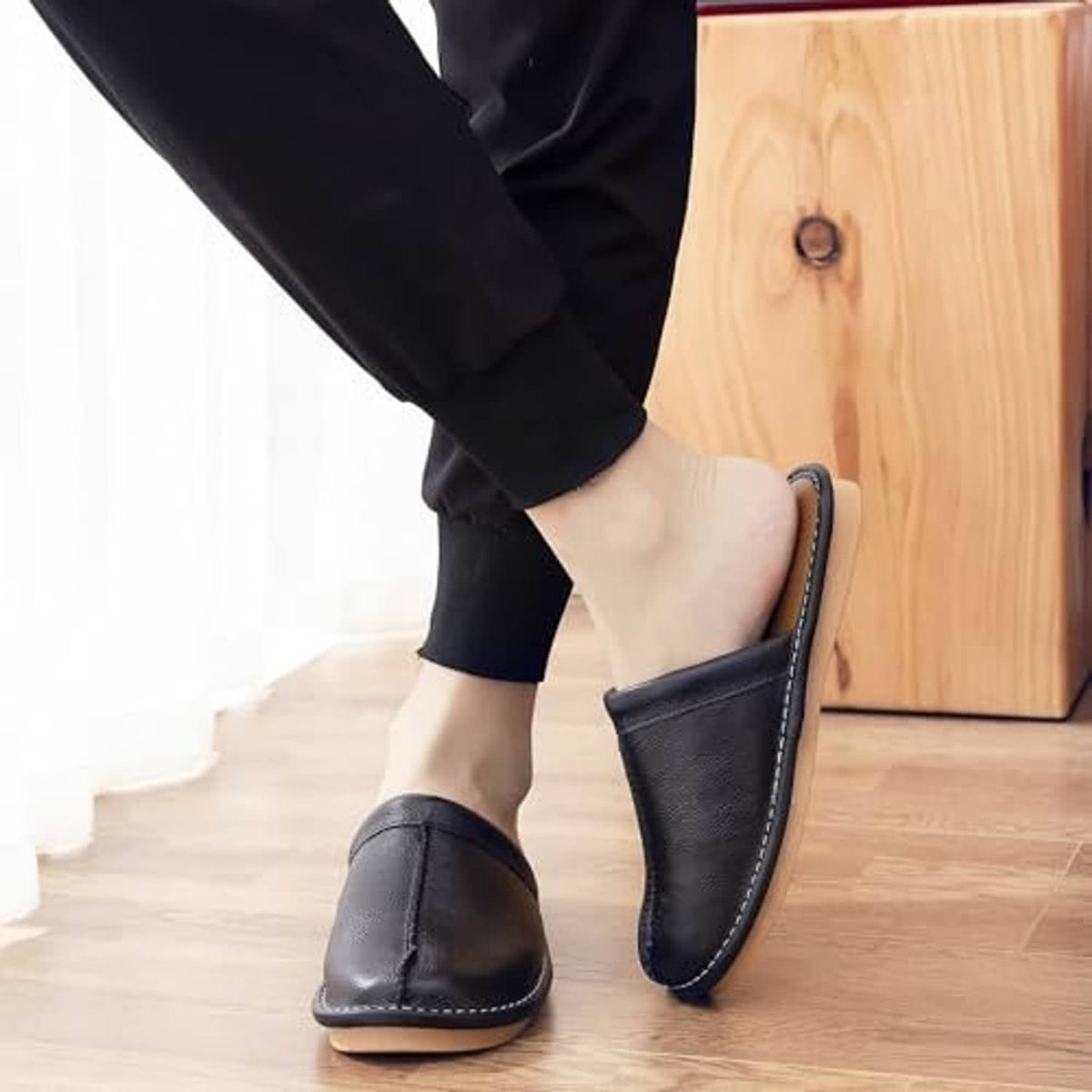 Black leather slip-on slippers with stitched edges shown on a light wood floor