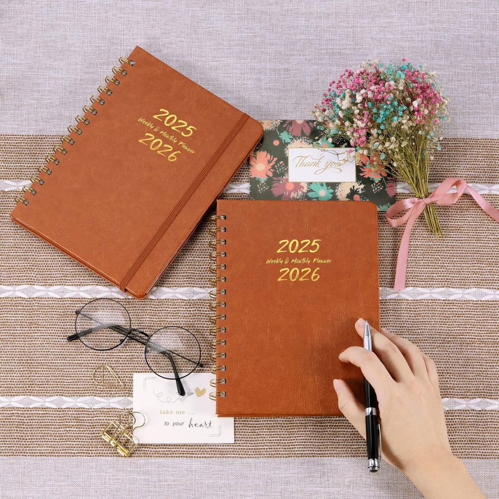 Two brown twin-wire planners with gold 2025 and 2026 covers, one being written on.