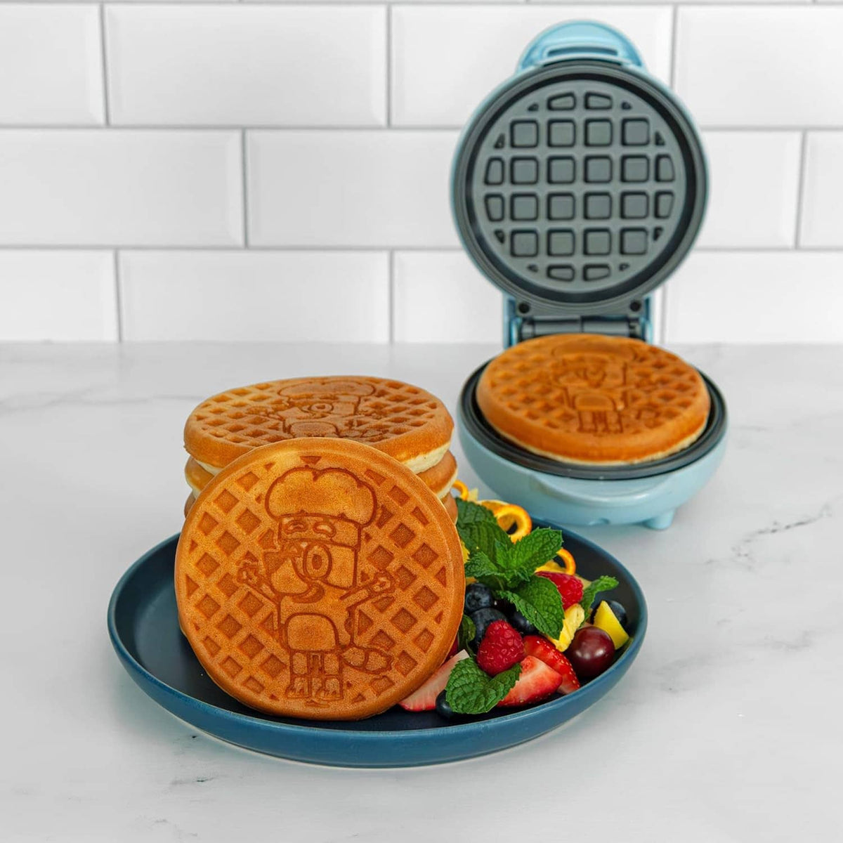 Light blue mini waffle maker open with two golden waffles and berry garnish on plate