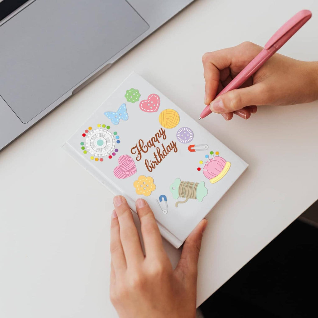 Hands using a pen on a sewing-themed card with cutout icons and tools