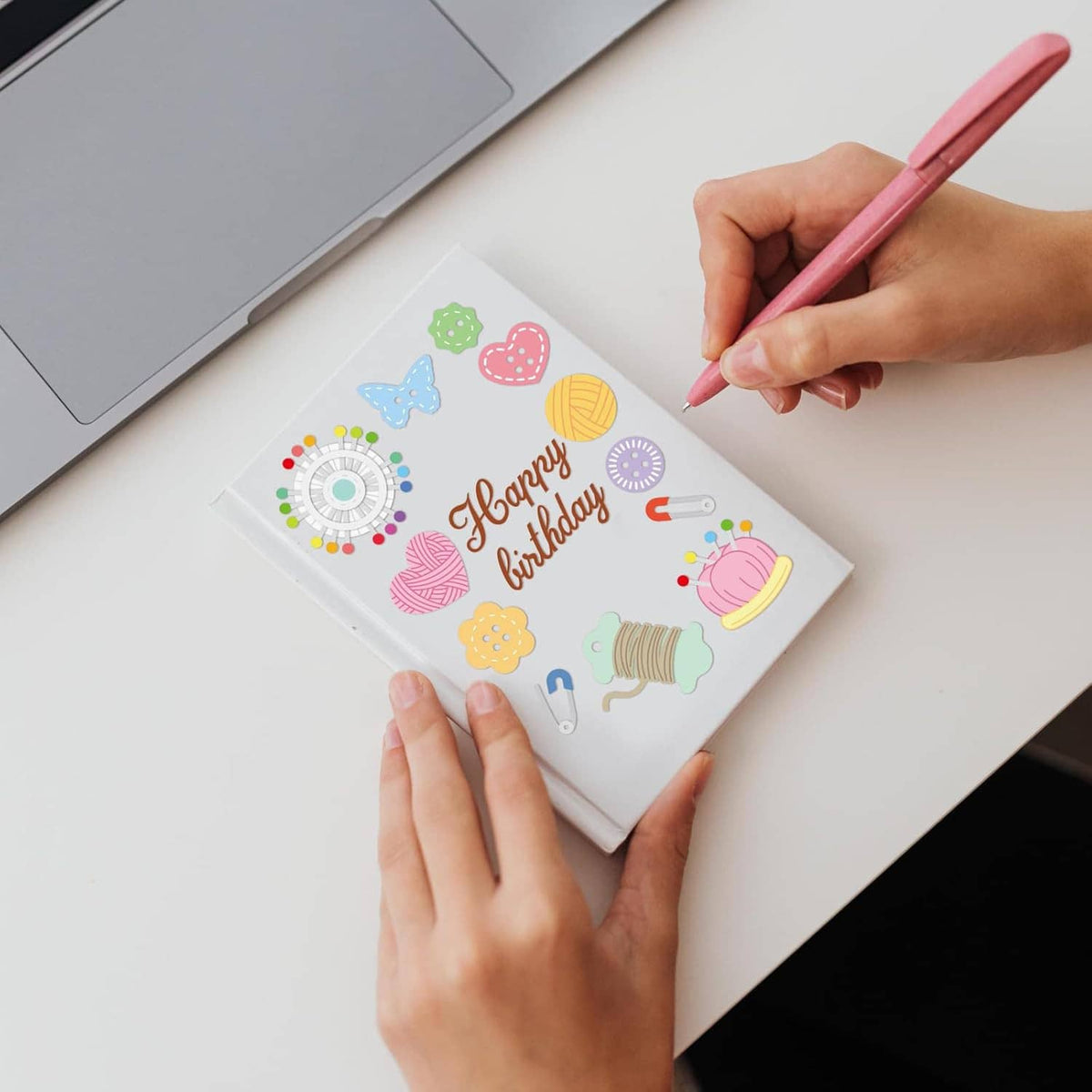 Hands using a pen on a sewing-themed card with cutout icons and tools