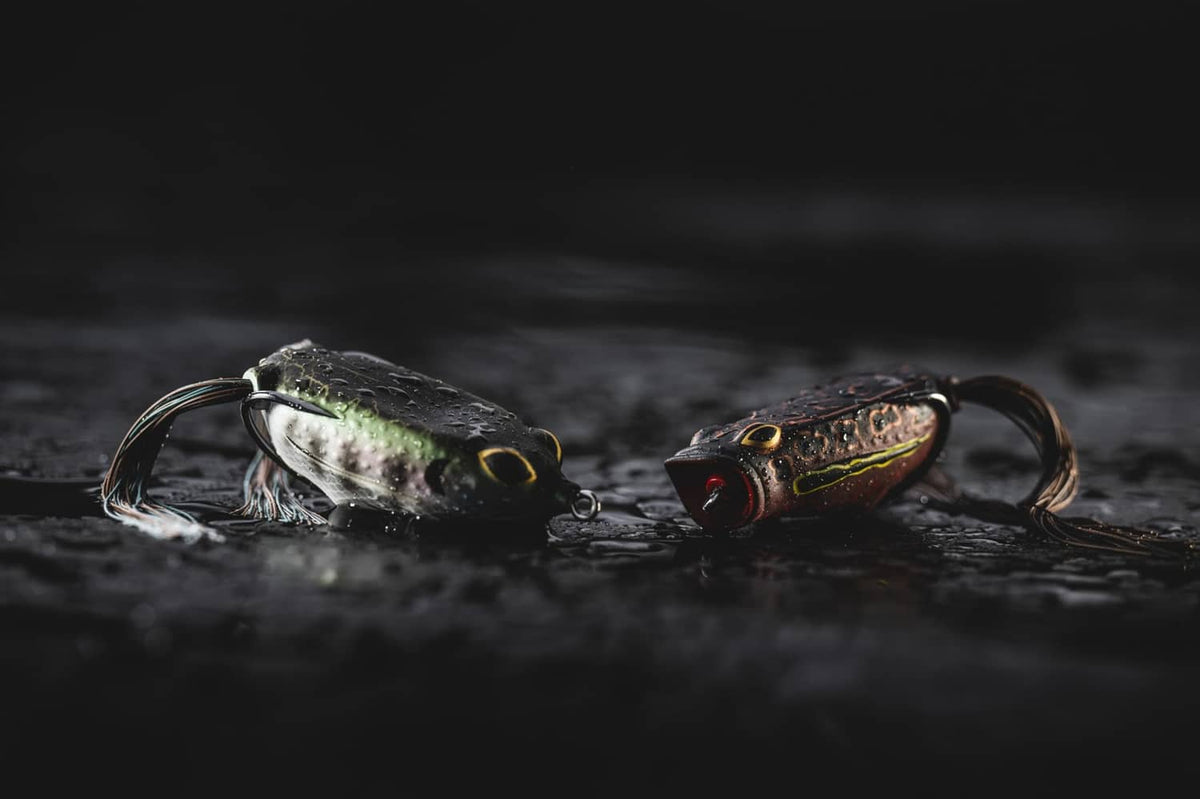 Two frog popping lures in water, dark camo bodies with yellow skirts