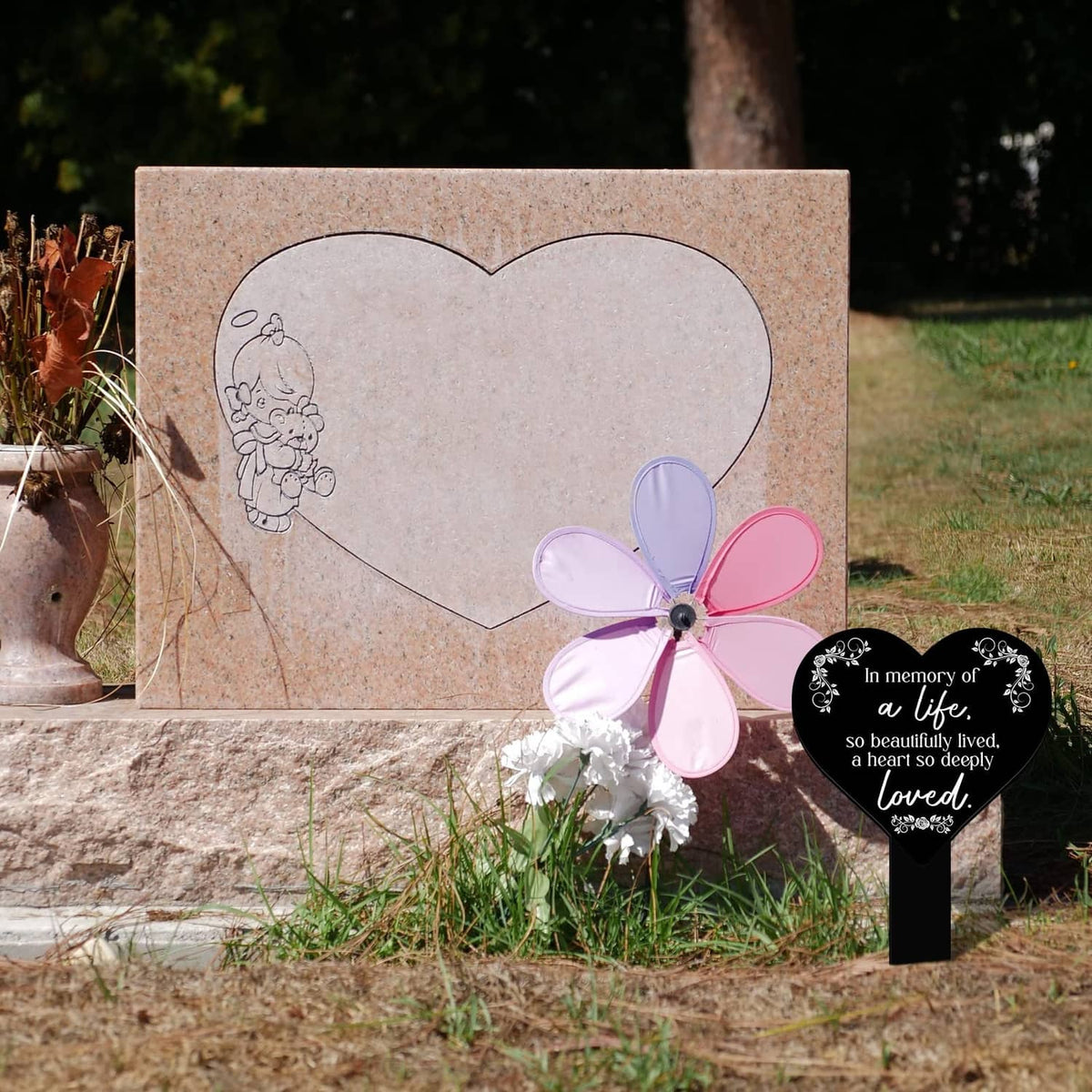 Heart-shaped black memorial plaque with white verse on stake beside a grave headstone.
