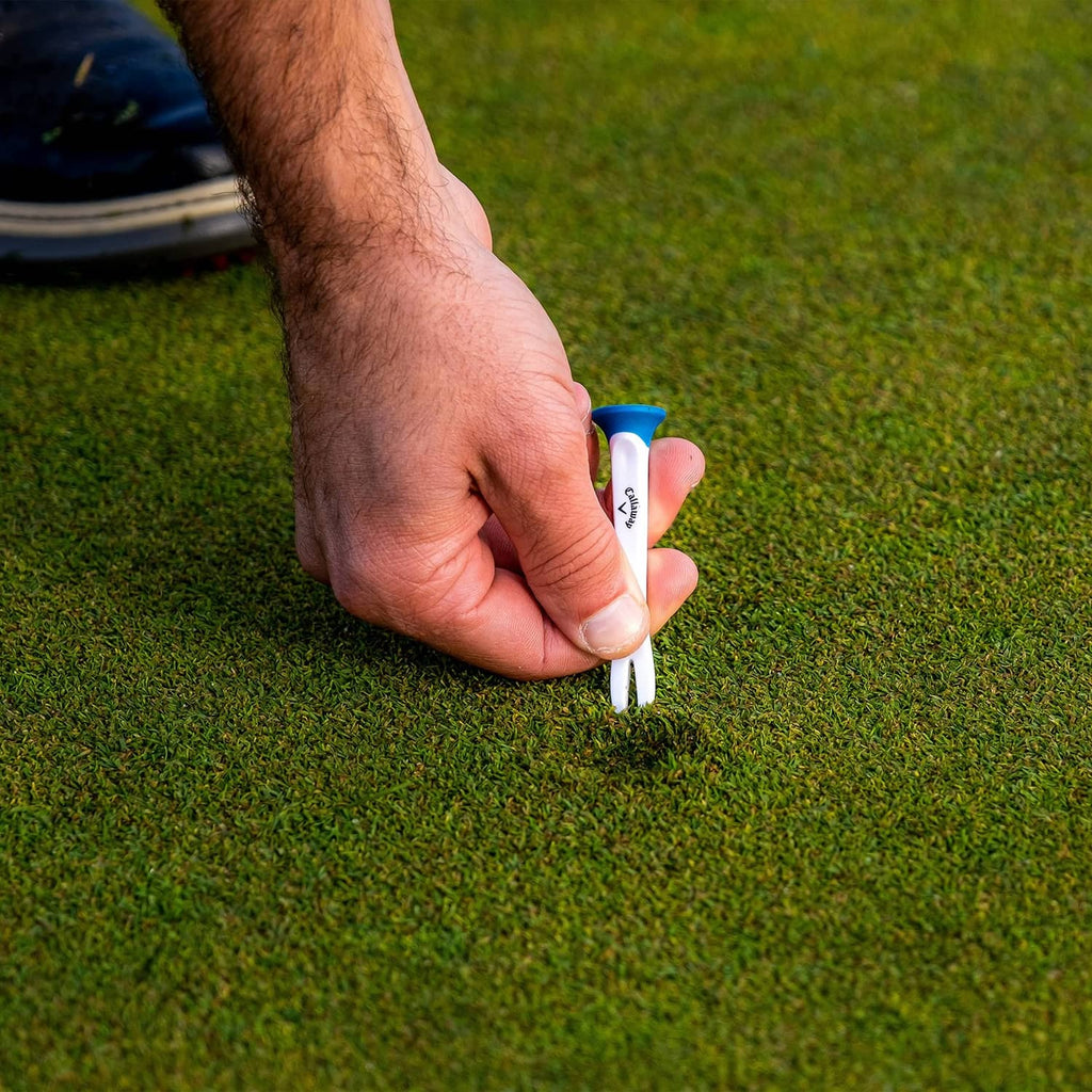 Hand pressing a white plastic golf tee with blue top into the turf
