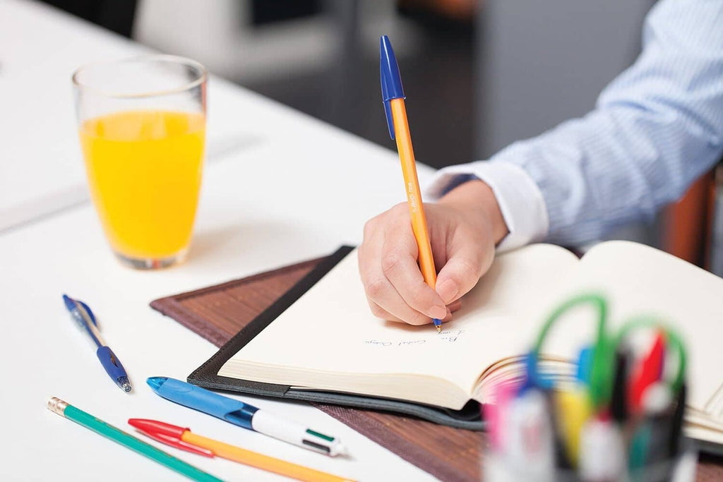 Person writing in notebook with an orange pen and blue tip on desk