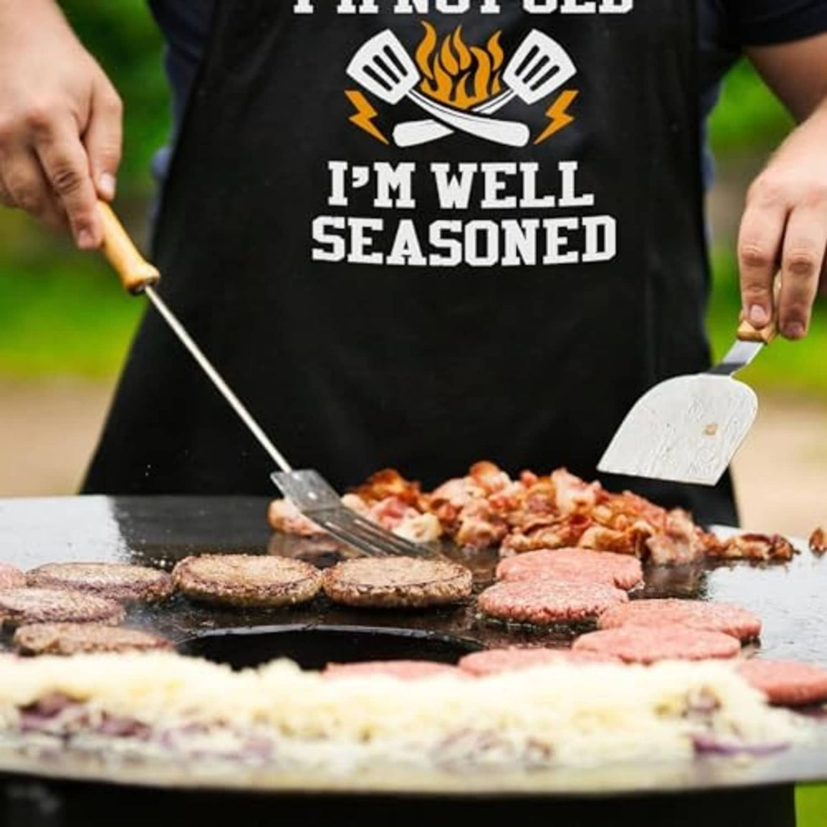 Black grilling apron with partial 'I'm well seasoned' print while cooking on a grill