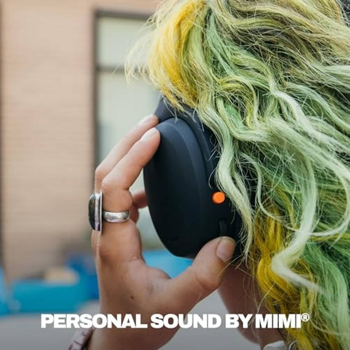 Black over-ear wireless headphones held to a person’s ear, long green-tipped hair visible