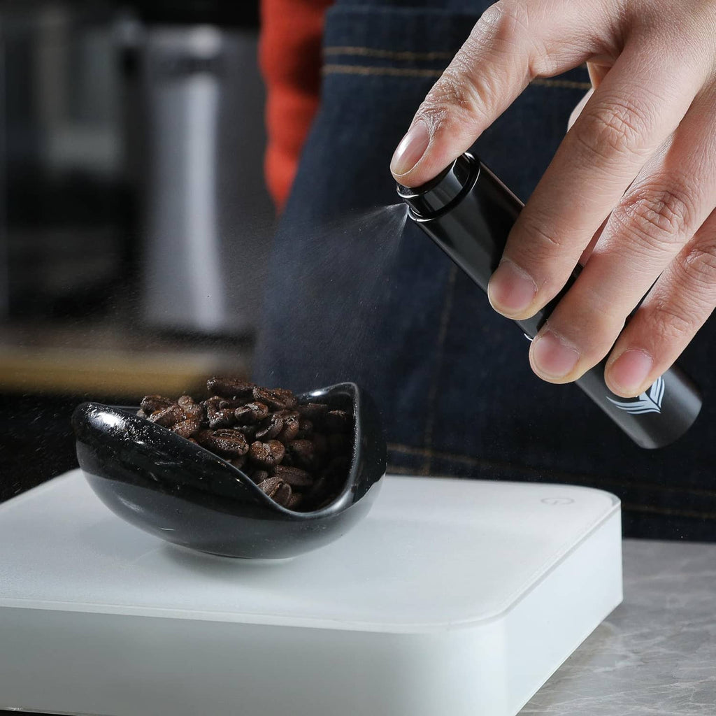 Hand spritzing coffee beans from a black RDT sprayer over a dosing cup