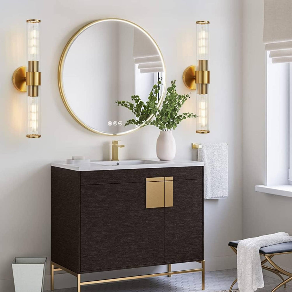 Gold vanity wall light 2-pack with clear glass tubes and round backplates near sink mirror.