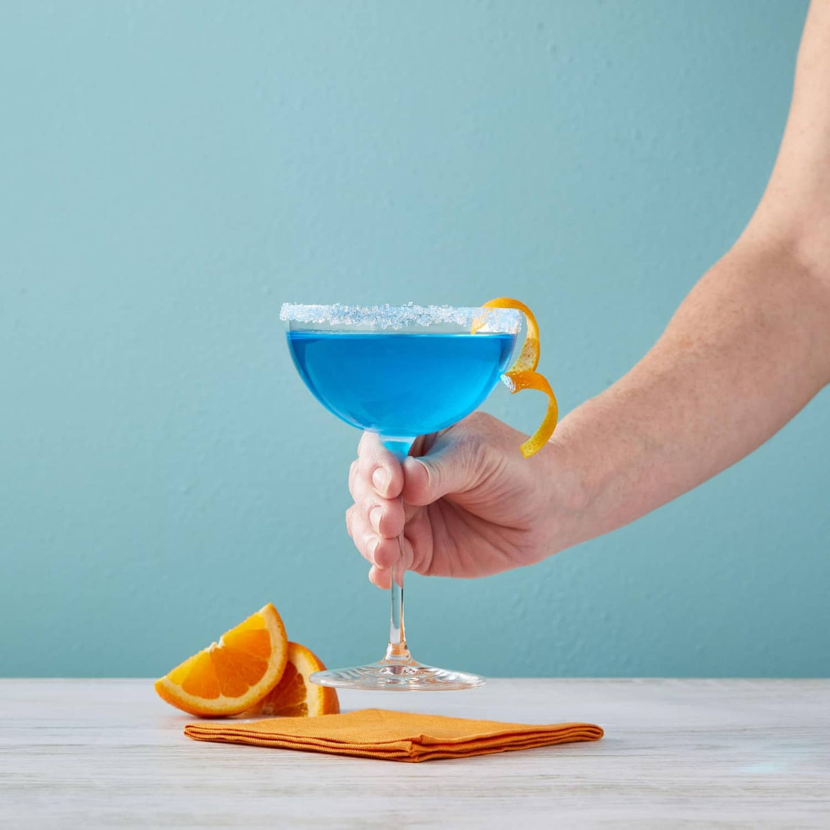 Hand holding blue cocktail in a stem glass with orange slice garnish and bottle