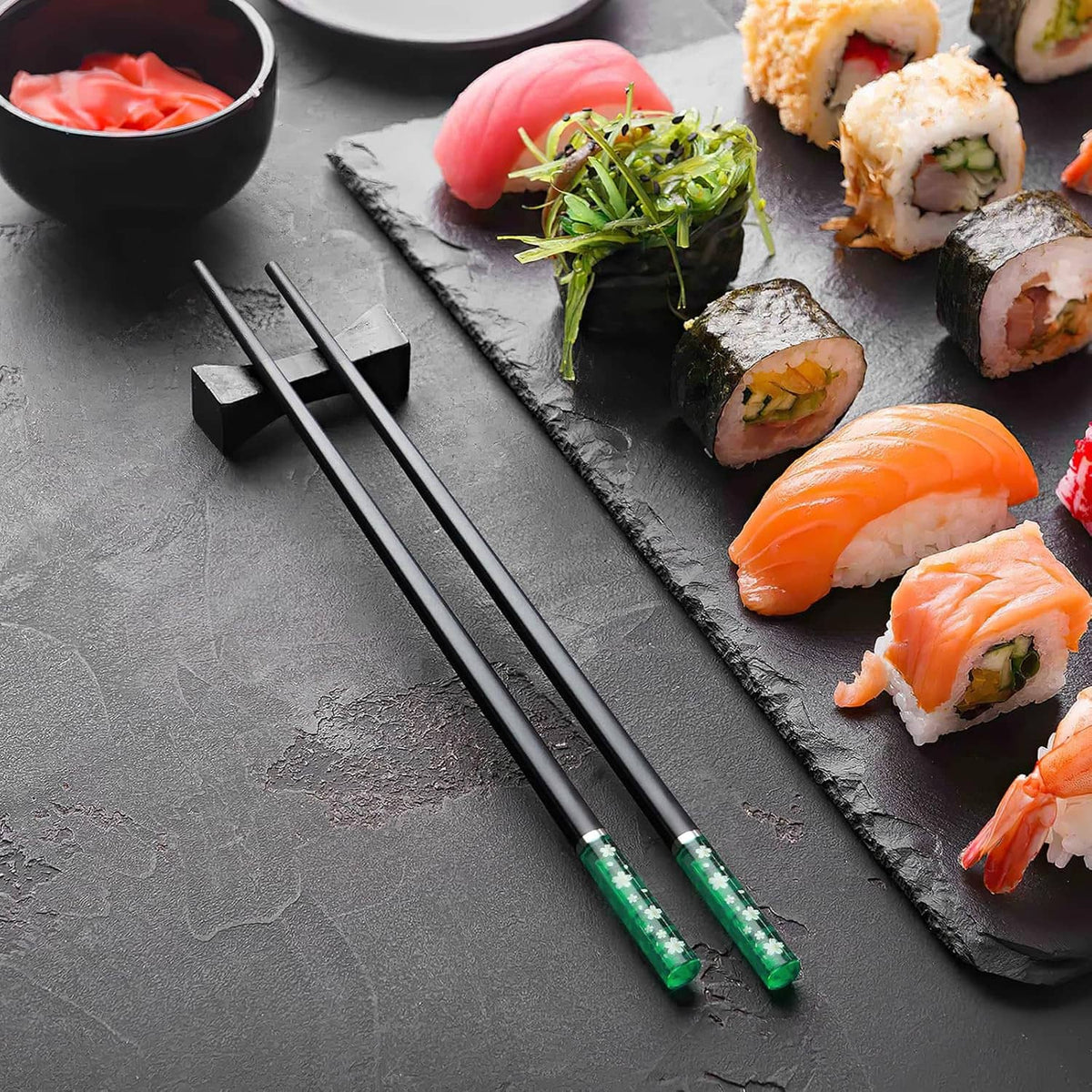 Chopsticks and sushi rolls on a slate serving board, with green-sleeved chopsticks