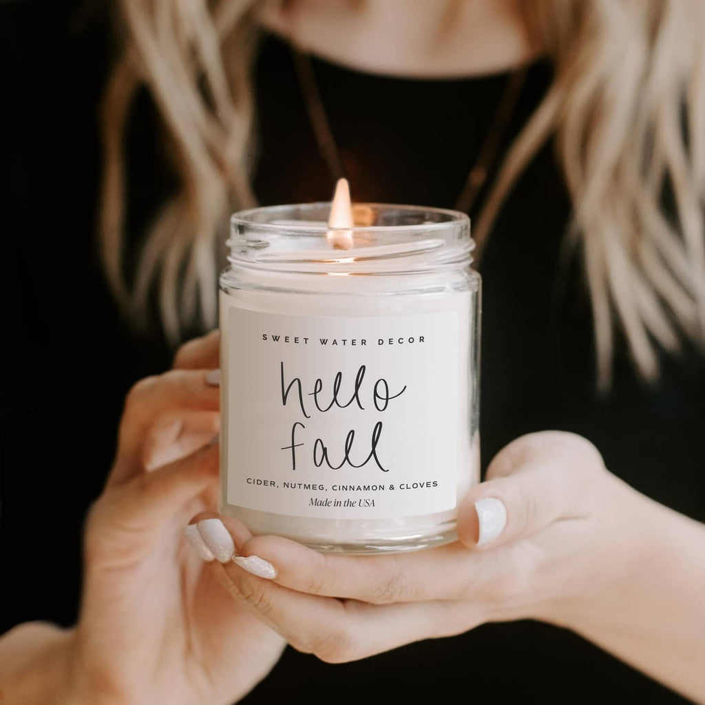 Hand holding a lit Hello Fall jar candle with cider, nutmeg, cinnamon, and cloves label