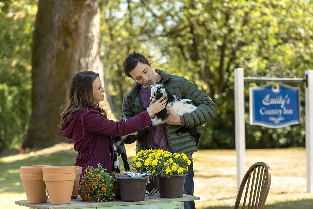 Film still of a couple interacting with a white-and-black dog outdoors near flowers