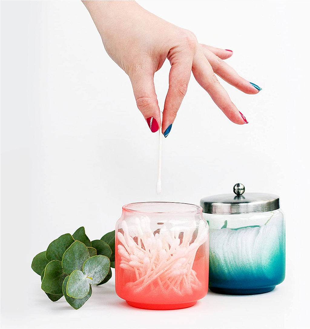 Hand holding a white cotton swab above small jars with pink and teal gel