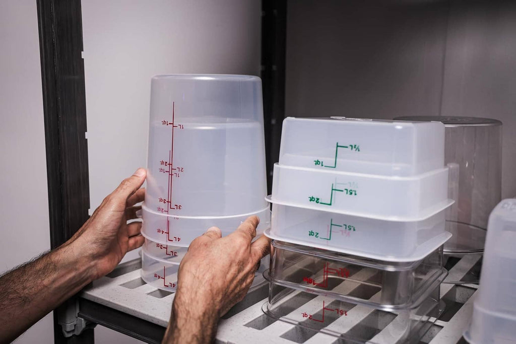 Stackable plastic food storage containers with lids, marked measurements, handled in an appliance area.