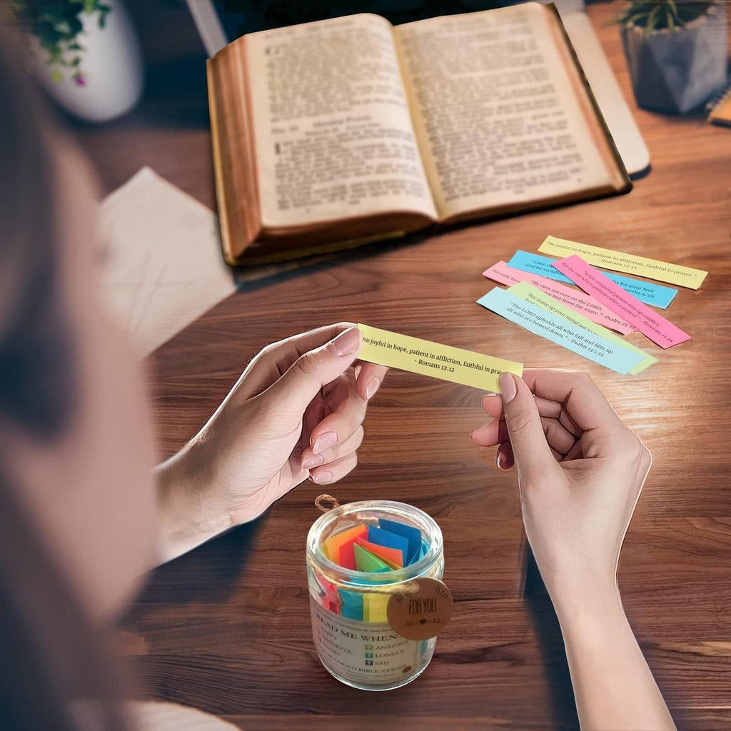 Hands placing colorful scripture slips into a small clear jar on a wooden table