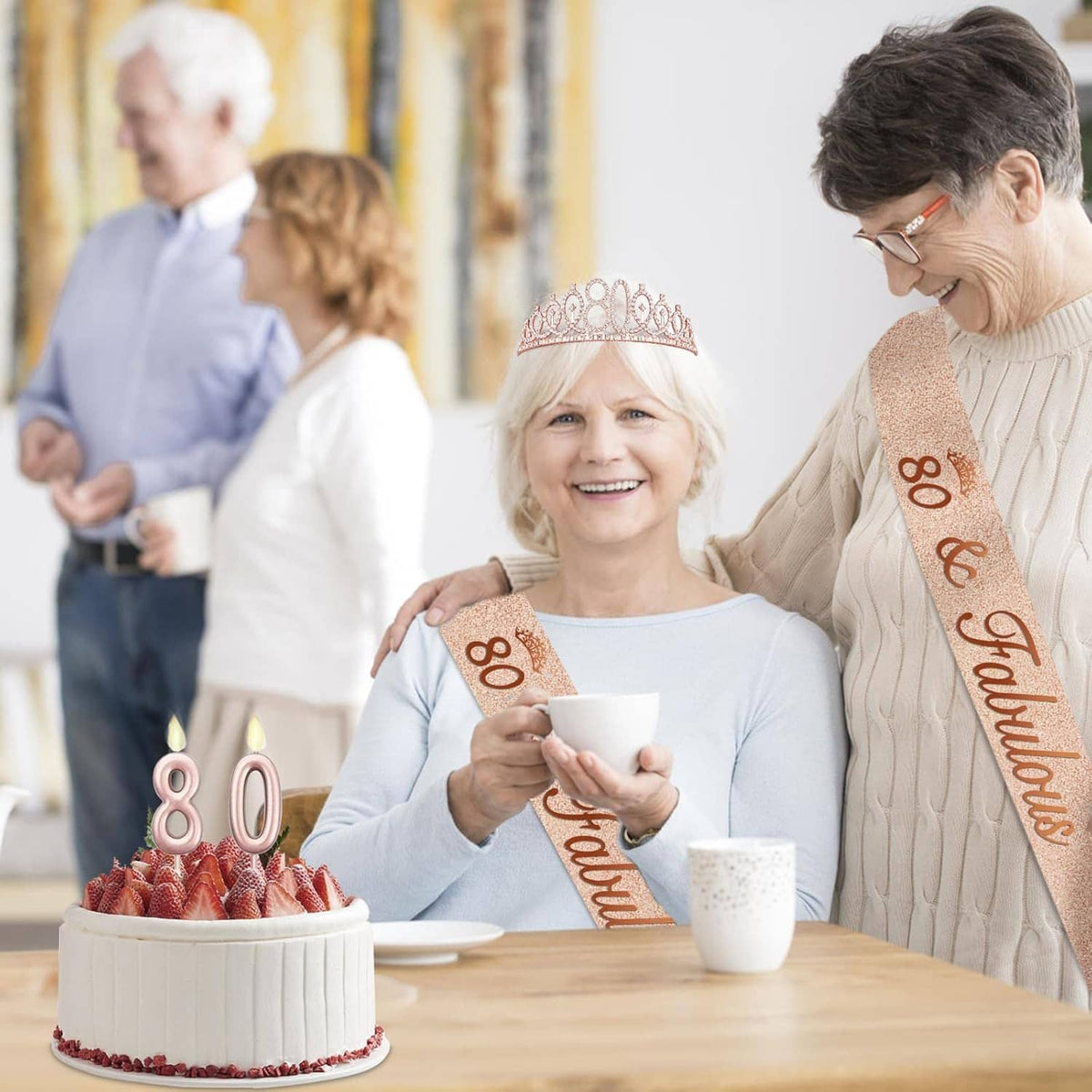 Elderly woman wearing rose-gold 80 tiara and sash beside a two-candle 80 cake