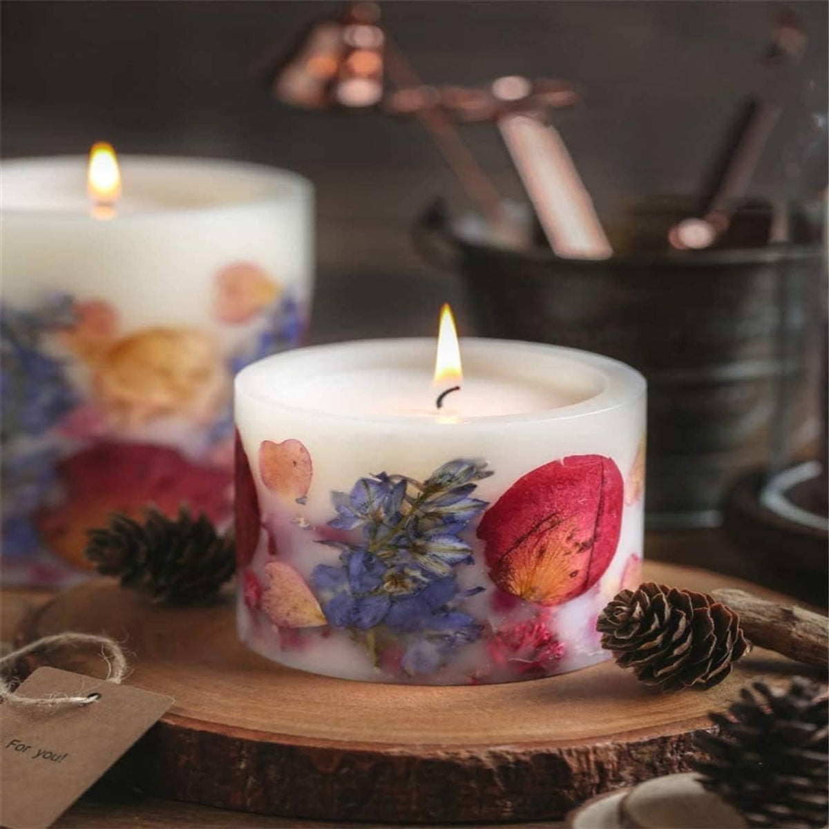White cylindrical pillar candle with embedded dried flowers and pinecones on a wooden base.