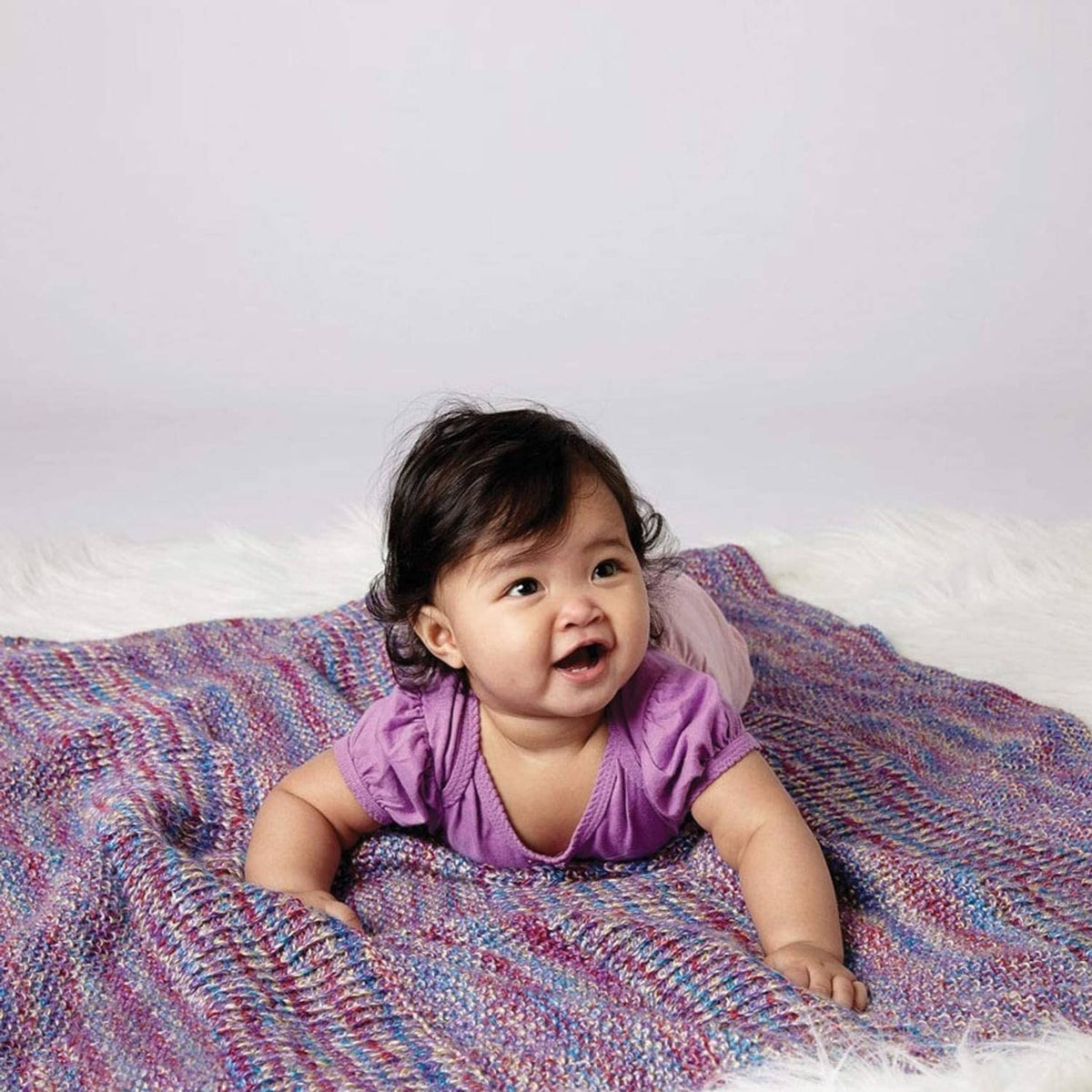 Baby lying on a multicolor knit blanket wearing a purple top