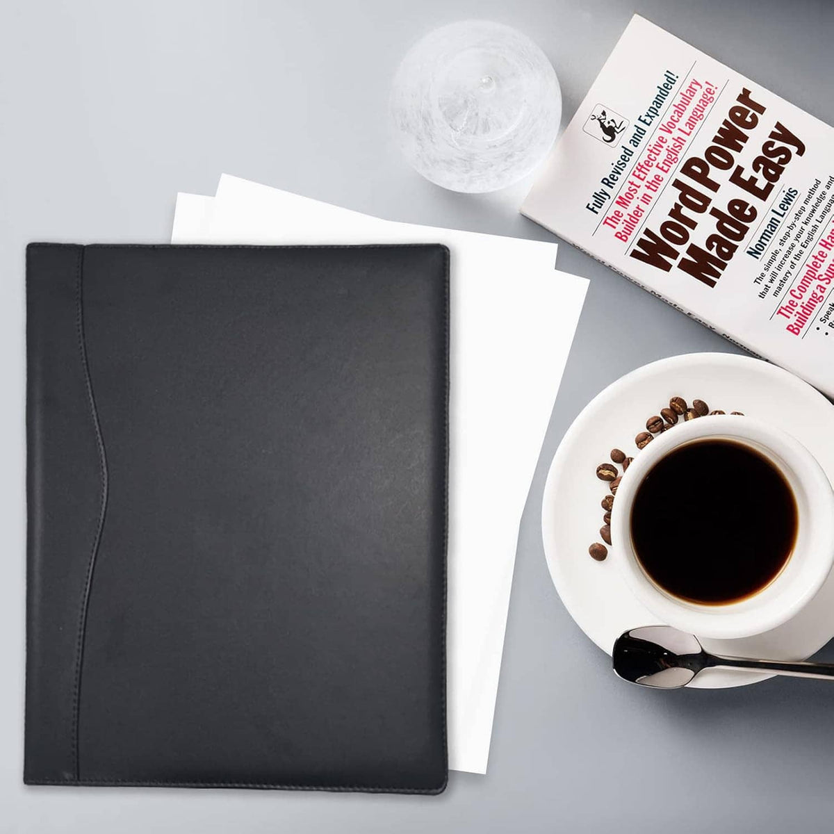 Black faux-leather padfolio with cover closed, laid flat with papers and coffee nearby