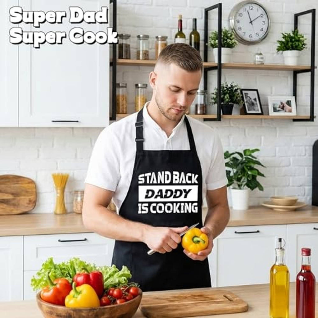 Black kitchen apron with front pocket and “STAND BACK DADDY IS COOKING” slogan print