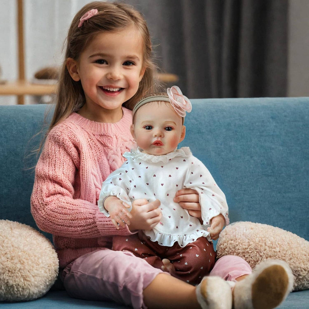 Realistic 18-inch reborn baby girl doll sitting with a girl in pink sweater, white heart outfit.