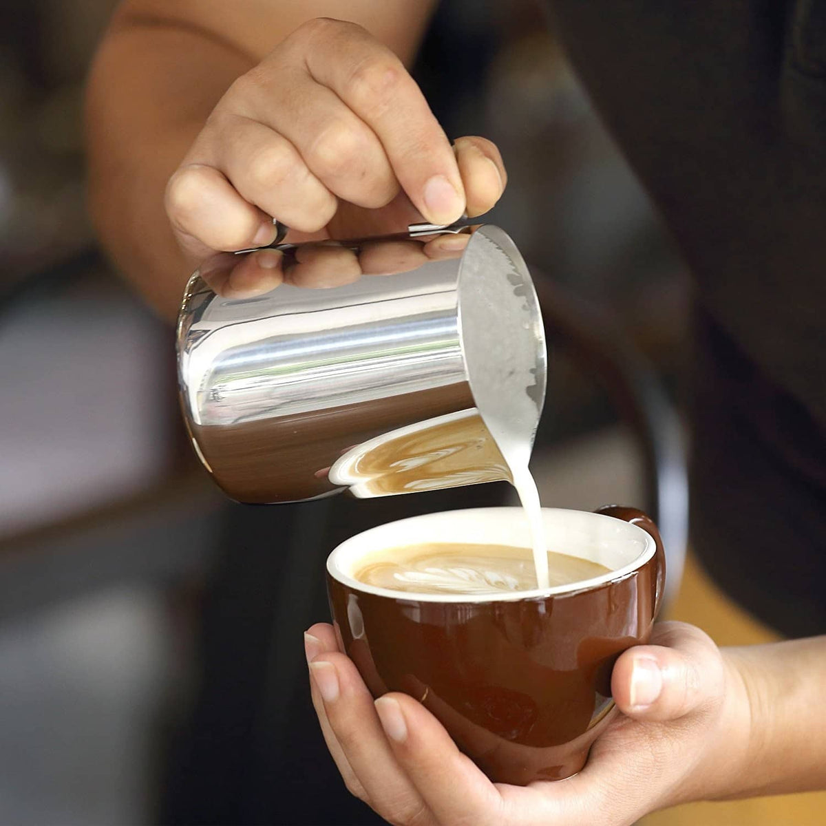 Stainless steel milk frothing pitcher being tilted to pour creamy coffee into a cup