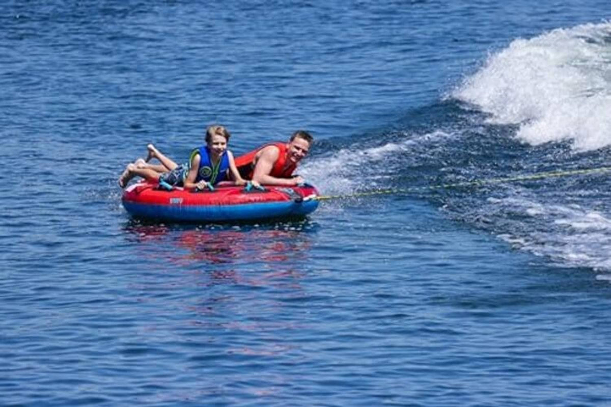 Red and blue two-person towable boat tube with two riders near the wake