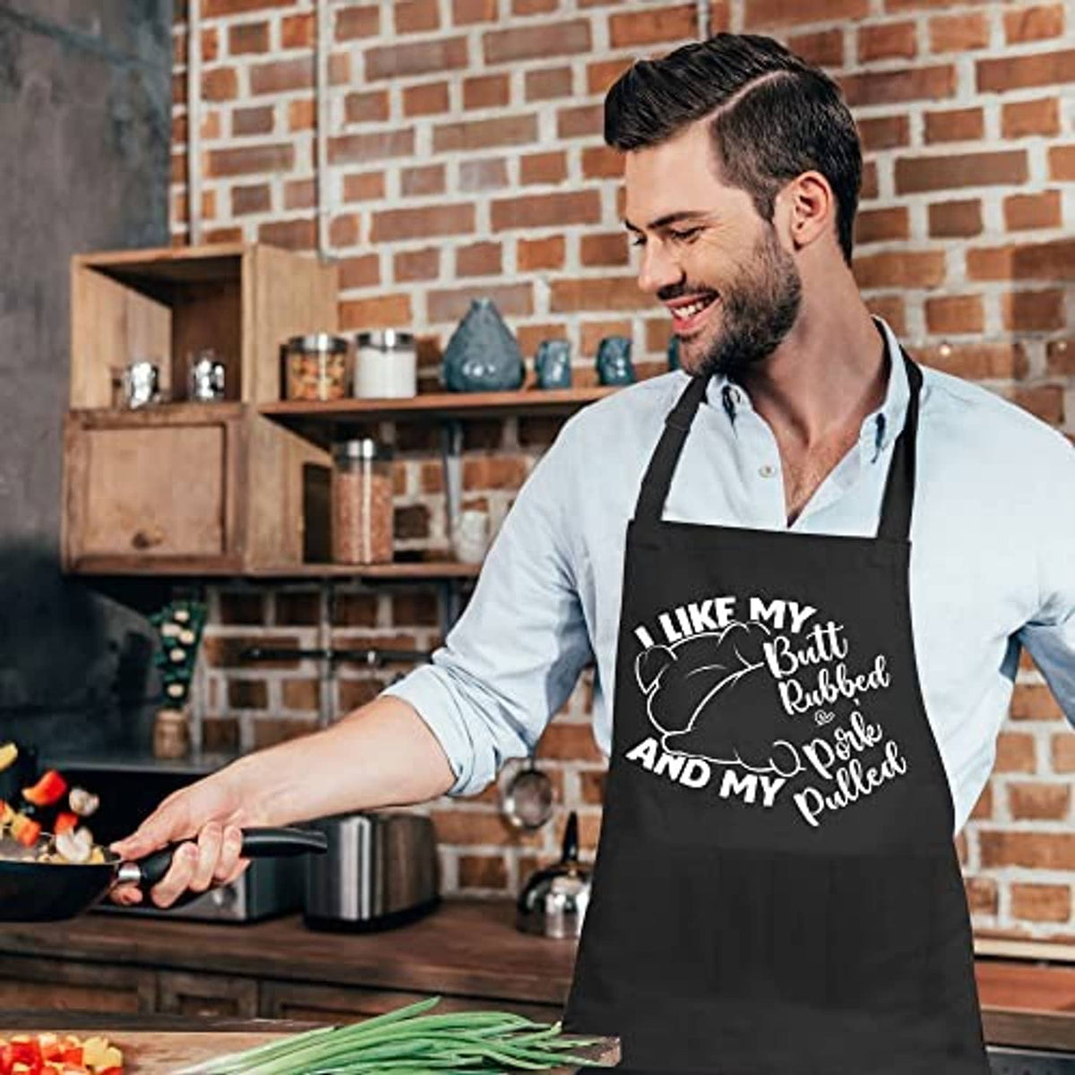 Black grilling apron on man by grill, front text and utensil graphics, bib-style design