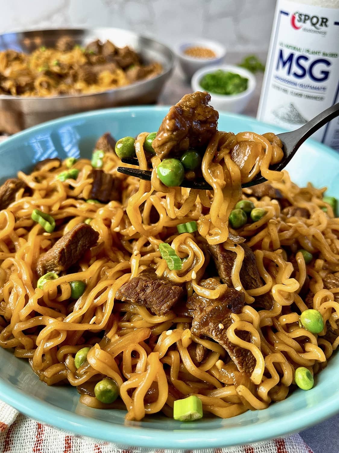 Bowl of sauced noodles with peas and meat pieces, with MSG seasoning jar in view.