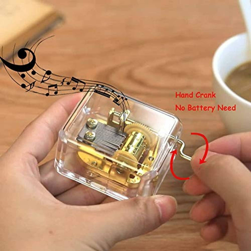 Clear hand-crank music box held in hand, gold mechanism visible through the transparent case.