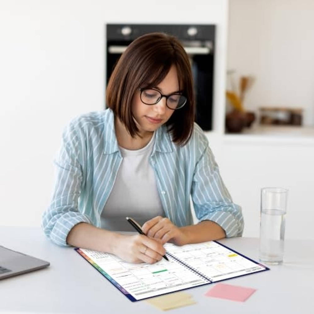 Person writing in a blue 2027 planner at a desk with scattered notes and a cup.