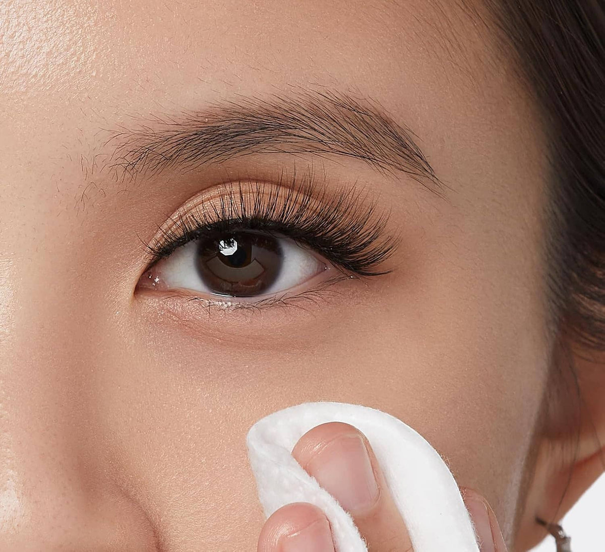 Eye with white cotton pad applying false eyelash cleanser along upper lashes