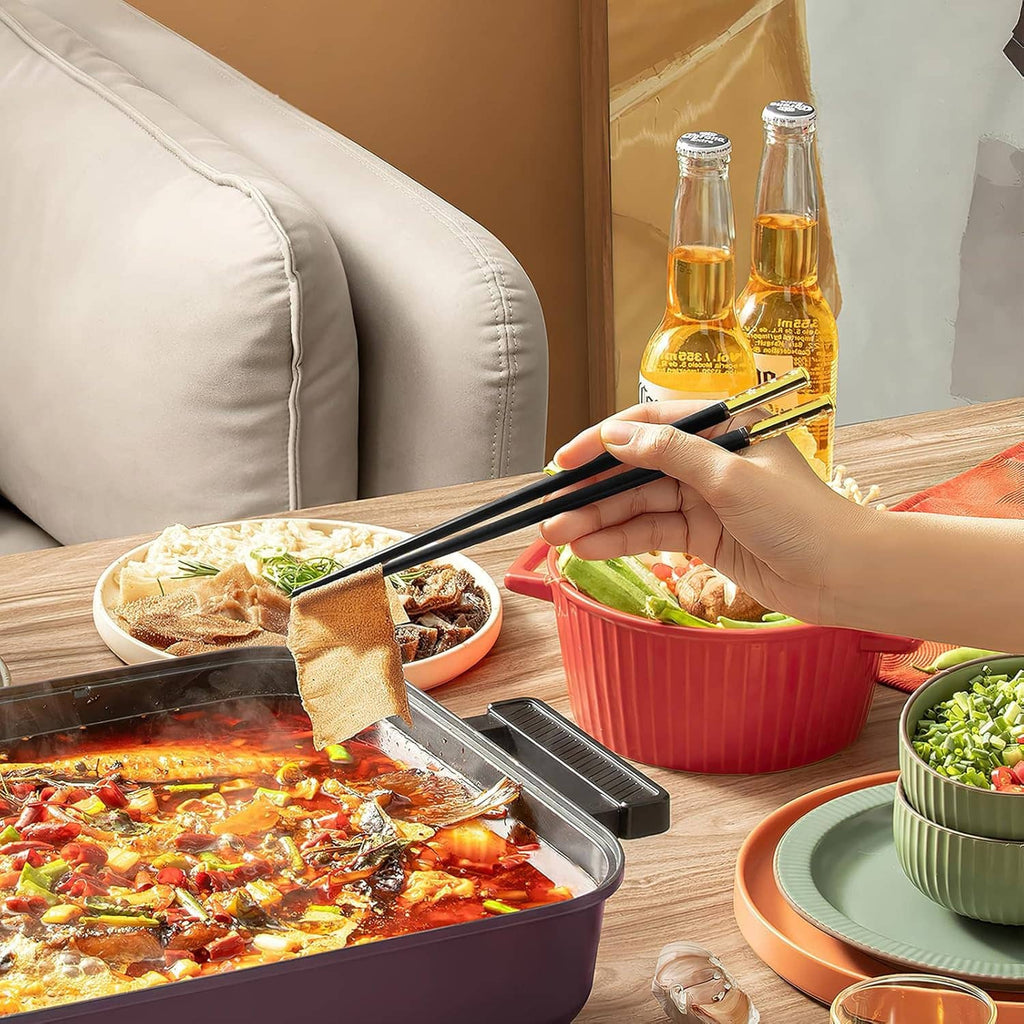 Hand holding black chopsticks over casserole and red bowl on a dining table