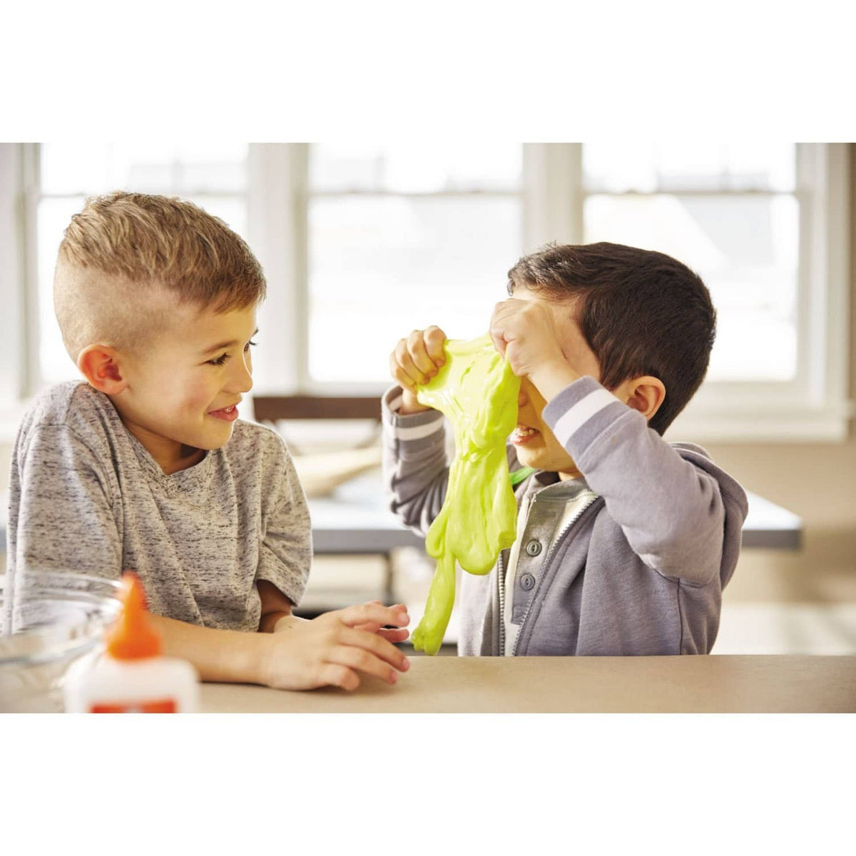 Kids pulling apart bright green slime with a bottle of Elmer’s school glue on table