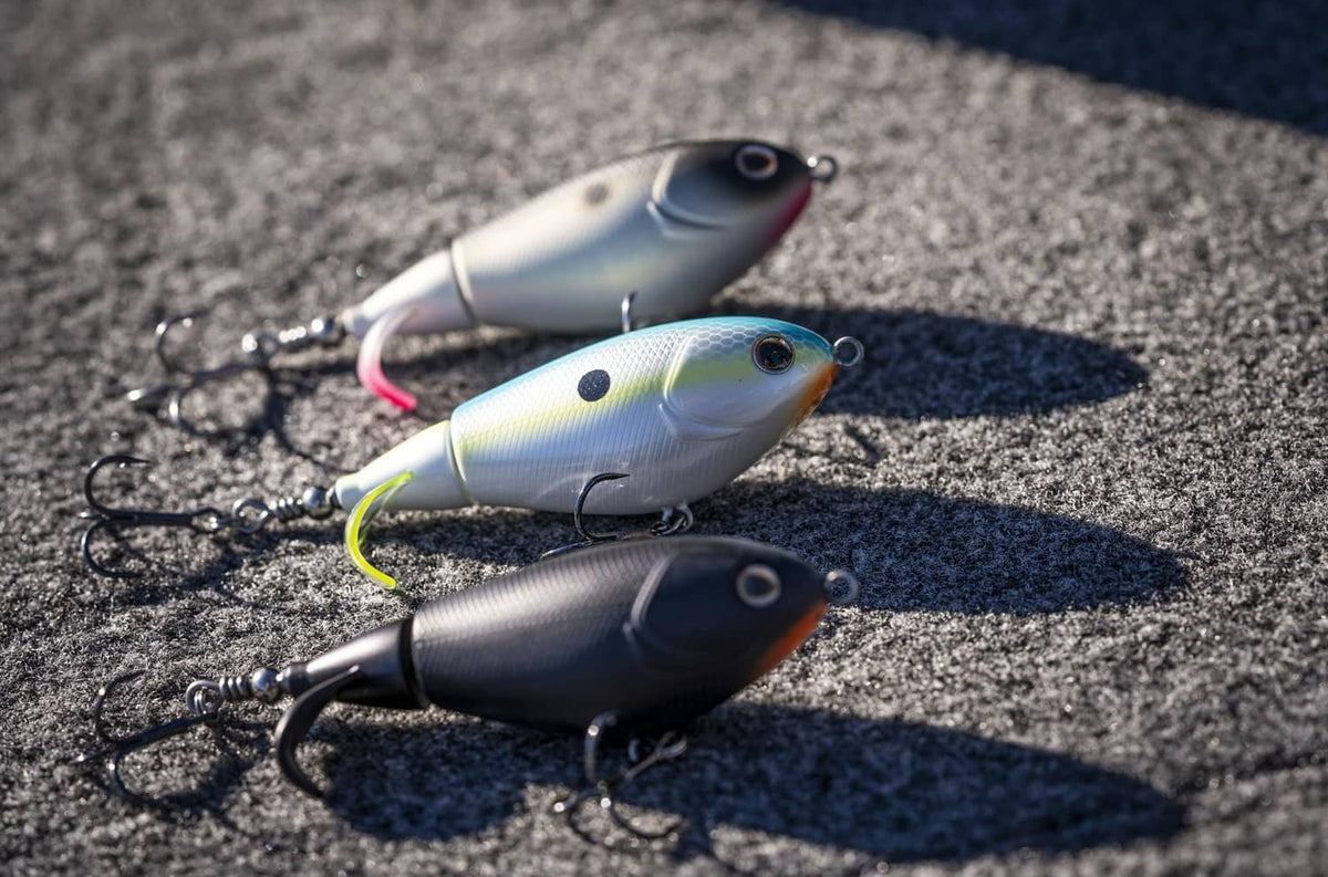 Three topwater lures on pavement in black, silver, and blue-gray with treble hooks