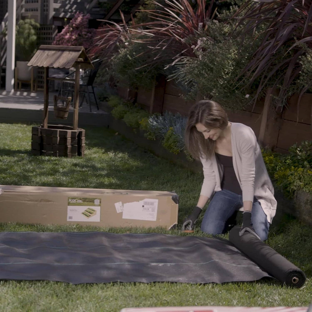 Woman unrolling a black weed barrier fabric sheet on grass while setting garden boxes.