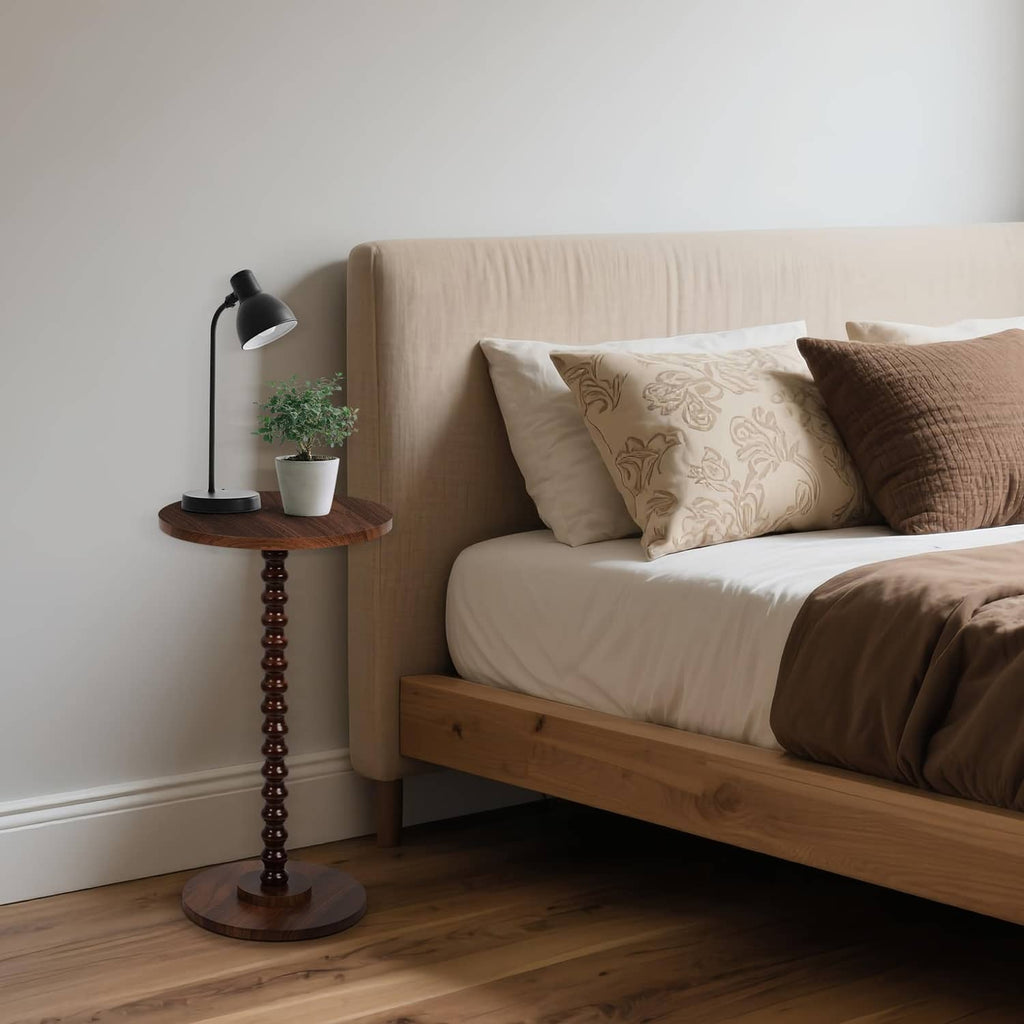 Rustic brown round side table with turned pedestal base beside bed and lamp