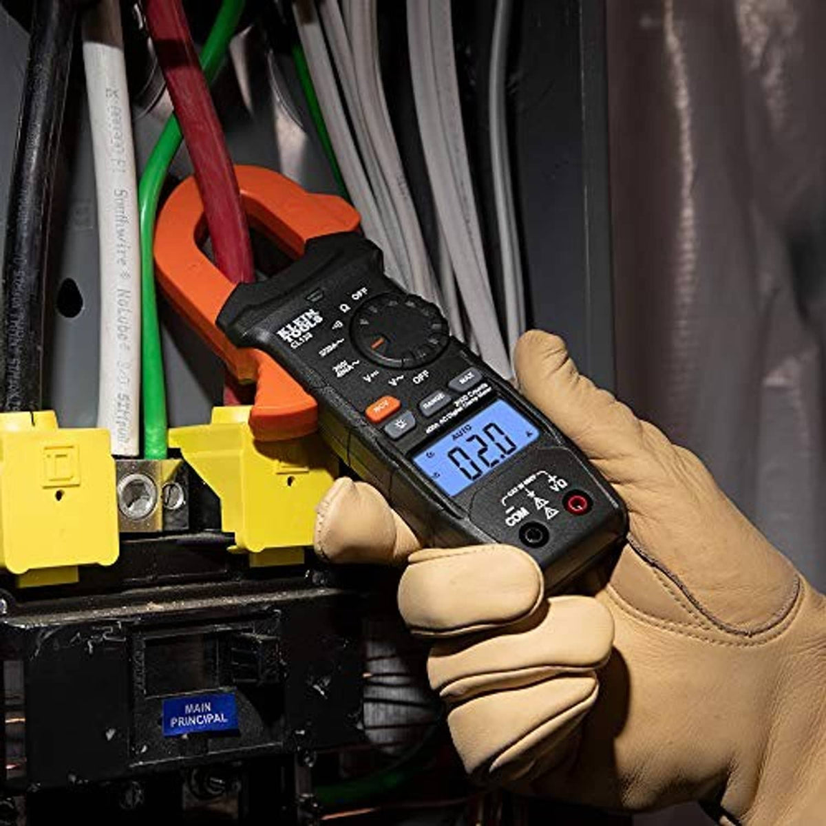 Digital clamp meter with blue screen held by gloved hands near bus wiring and breakers.