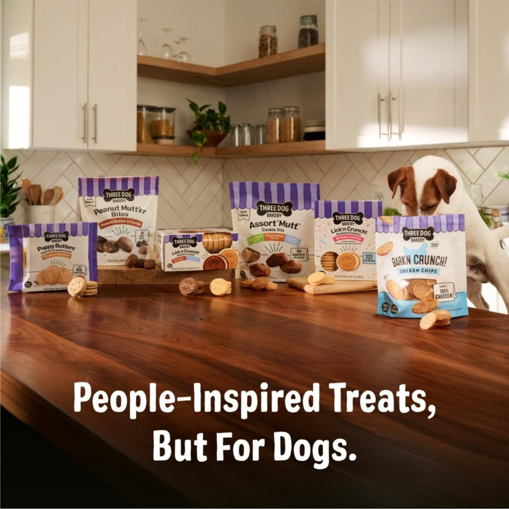 Assorted Three Dog Bakery treats packages on a table with dogs in the foreground