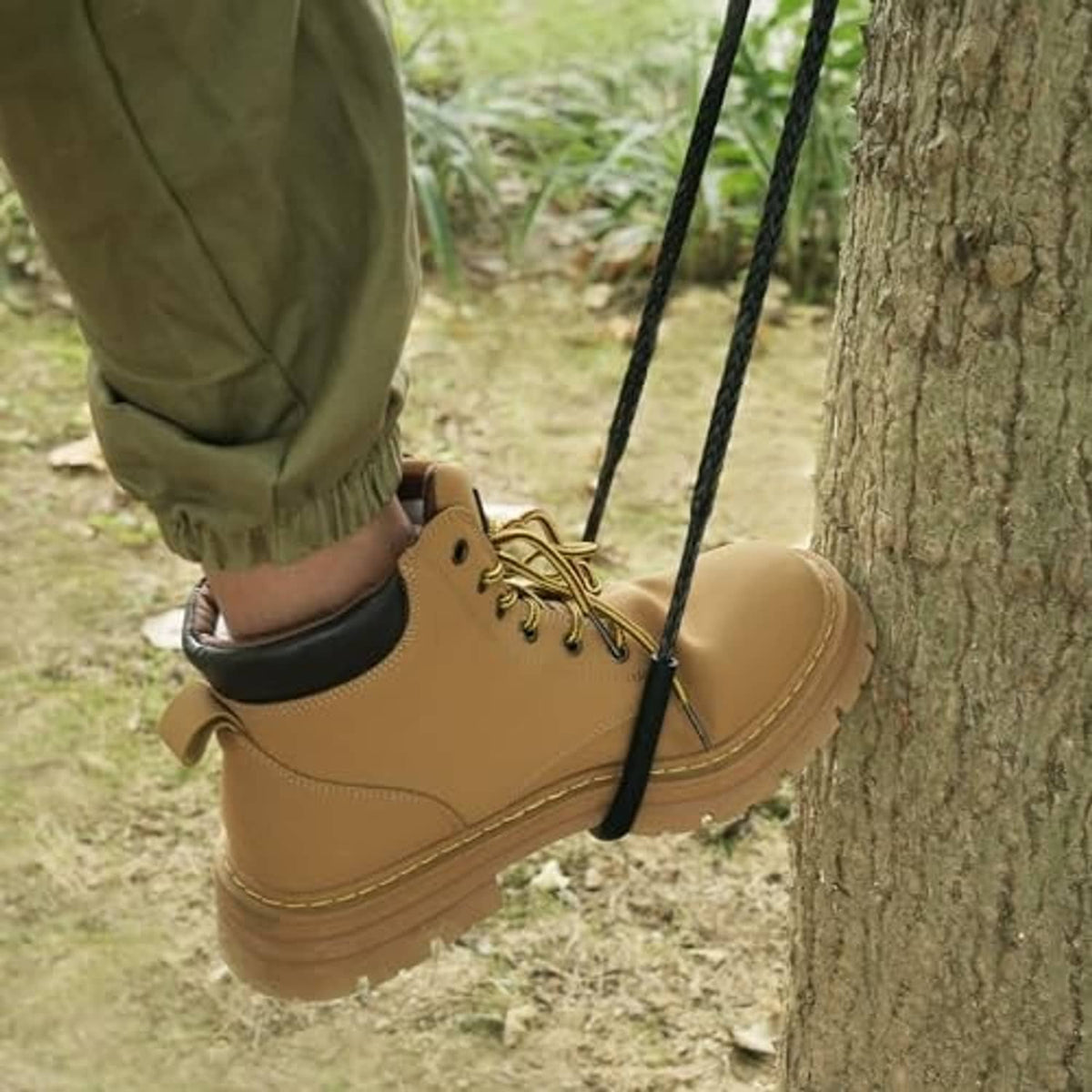 Tree climbing aider in use: black rope step next to a tan boot on the ground path.