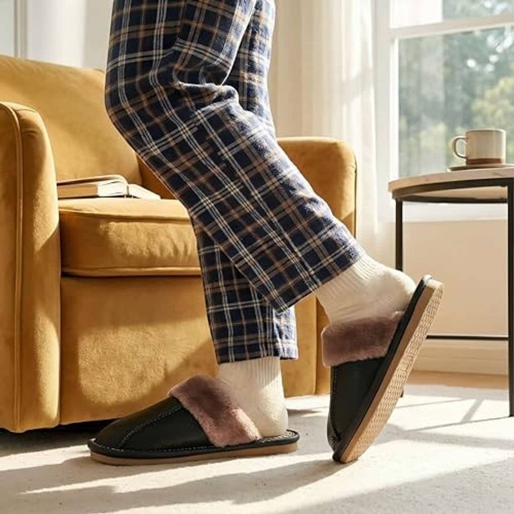 Unisex black leather slippers with plush taupe lining worn with socks by a sofa.