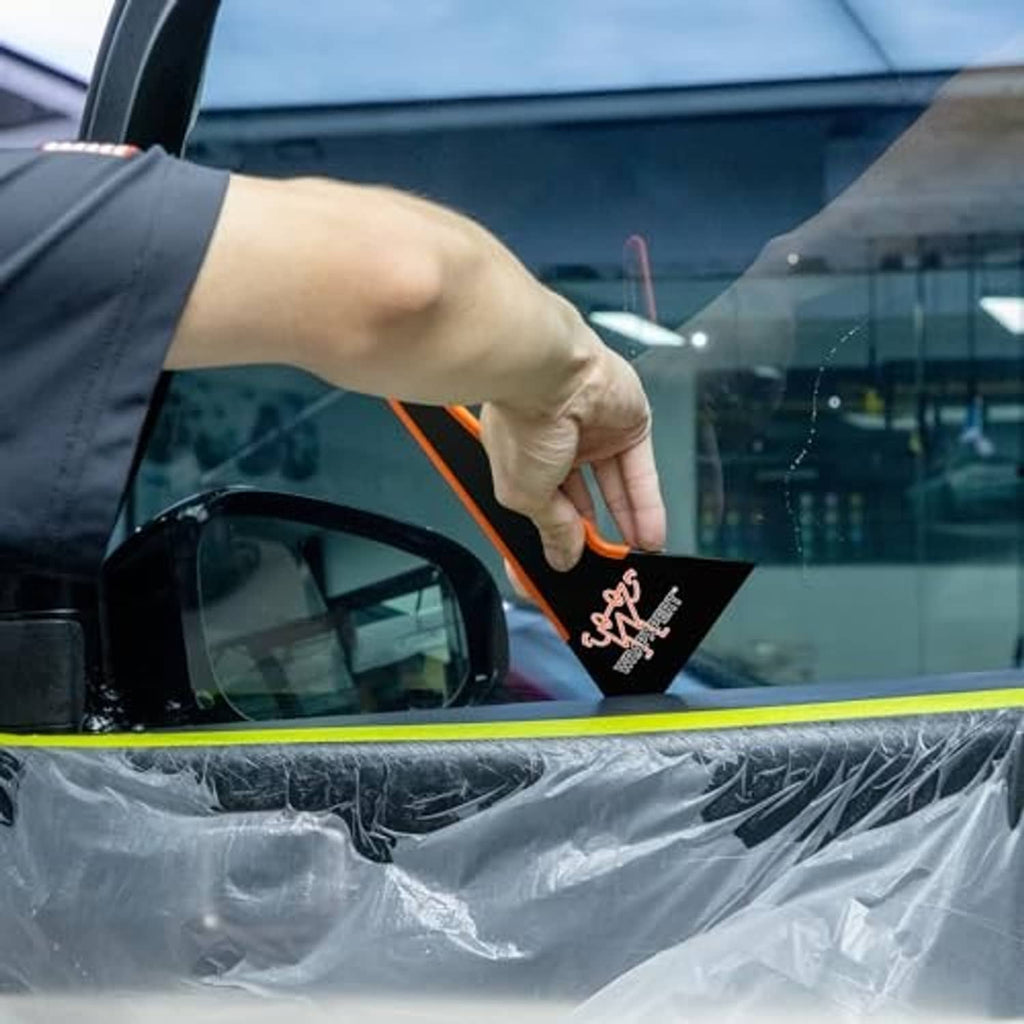 Hand using a black-and-orange window tint squeegee with triangular blade on a car window