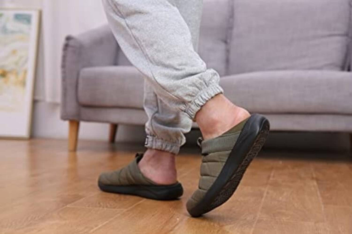 Army green slip-on house slippers on person’s feet with gray jogger pants