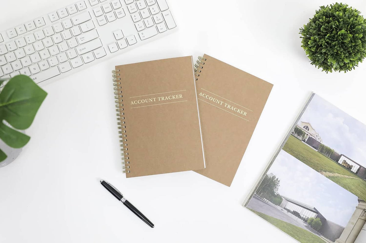 Two tan spiral-bound account tracker ledgers with gold spine and centered “ACCOUNT TRACKER”