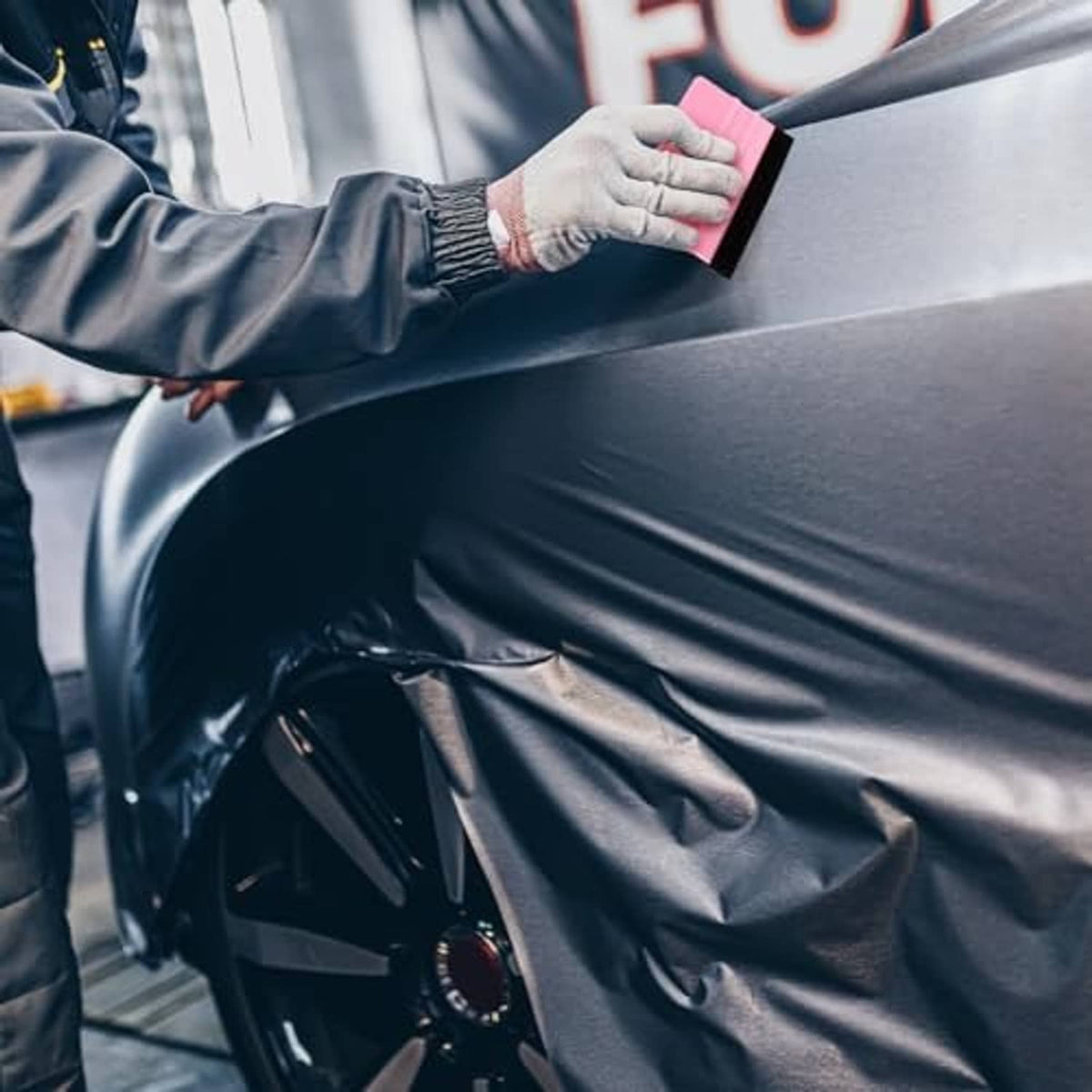 Hand using a pink felt-edge squeegee to smooth a vinyl wrap on a car panel