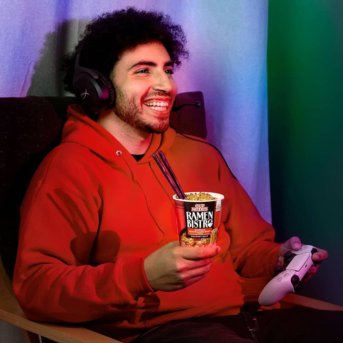 Red hoodie person holding Ramen Bistro noodle cup while using a controller