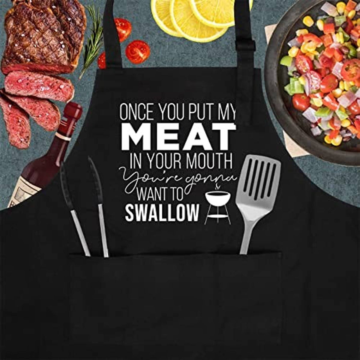 Black cooking apron with large "MEAT...SWALLOW" print and grill utensil icons.